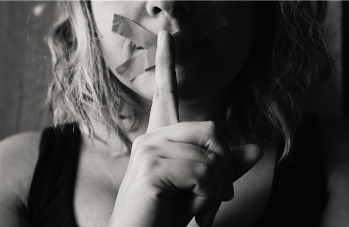 Photo of woman with duct tape criss-crossed over her mouth, holding her finger to it